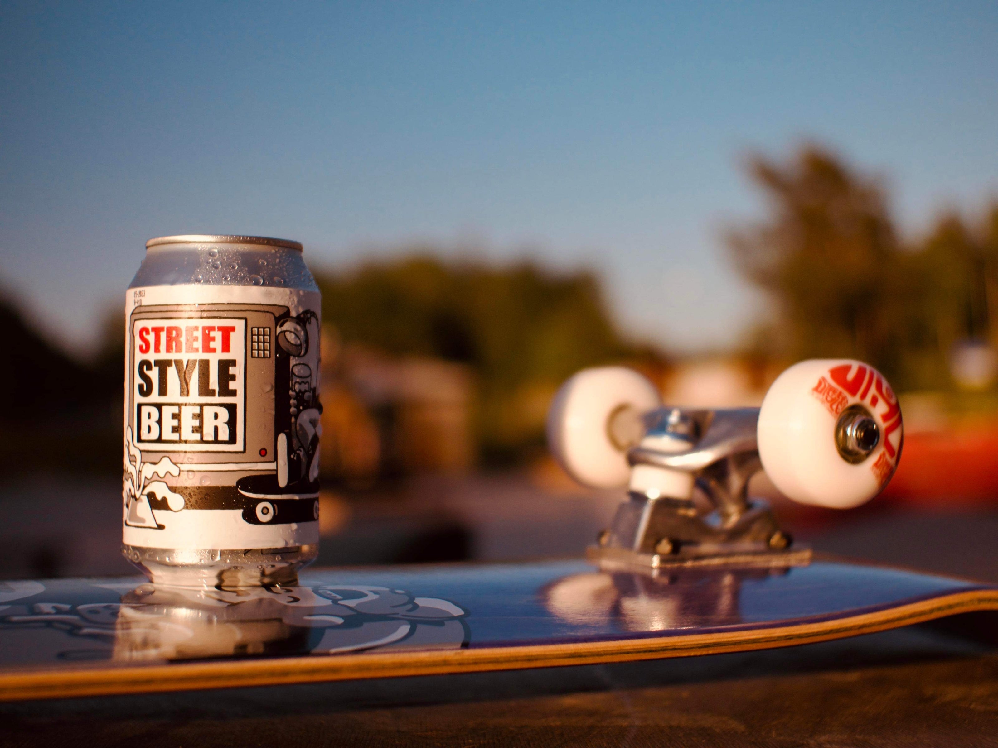 A can of "Street Style Beer" rests on a skateboard. The scene is set outdoors in warm, golden light, with a blurred background of trees. The skateboard is angled to showcase its deck and white wheels.