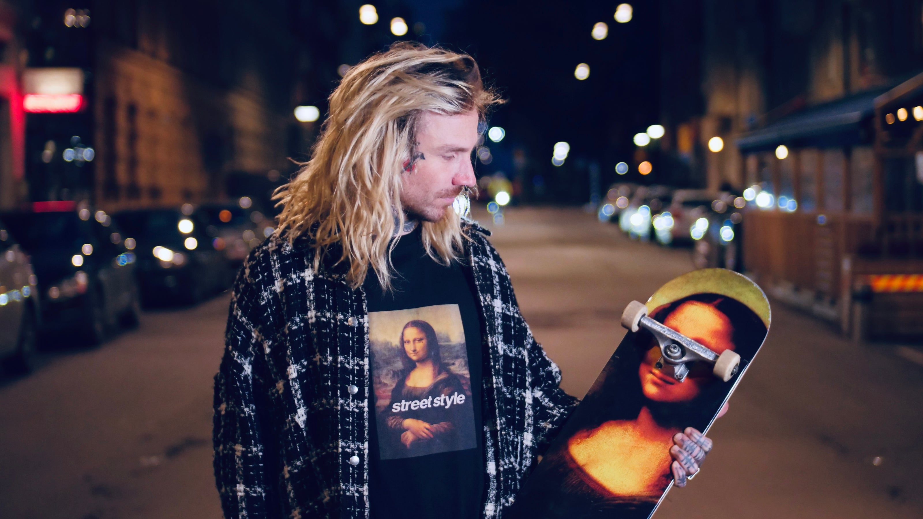 A person with long hair stands on a dimly lit street at night, holding a skateboard featuring the Mona Lisa. They are wearing a plaid shirt over a T-shirt with the Mona Lisa printed on it. Cars are parked along the street.